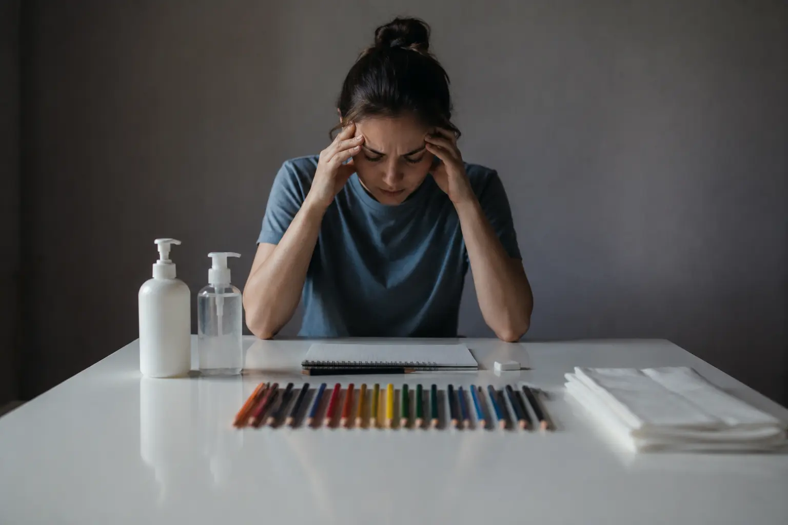 A stressed young woman sitting at a white table with sanitizer bottles, colored pencils, and tissues, symbolizing obsessive compulsive disorder (OCD), anxiety, and mental health struggles