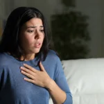 A woman experiencing a panic attack sits on a living room sofa with her hand on her chest, illustrating the symptoms of panic disorder in a realistic home setting 
