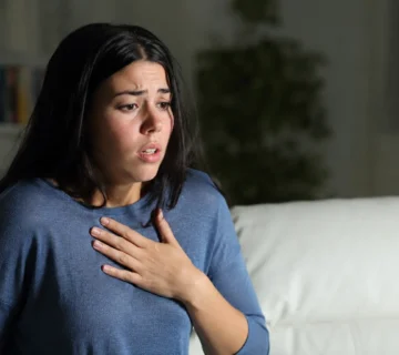 A woman experiencing a panic attack sits on a living room sofa with her hand on her chest, illustrating the symptoms of panic disorder in a realistic home setting 