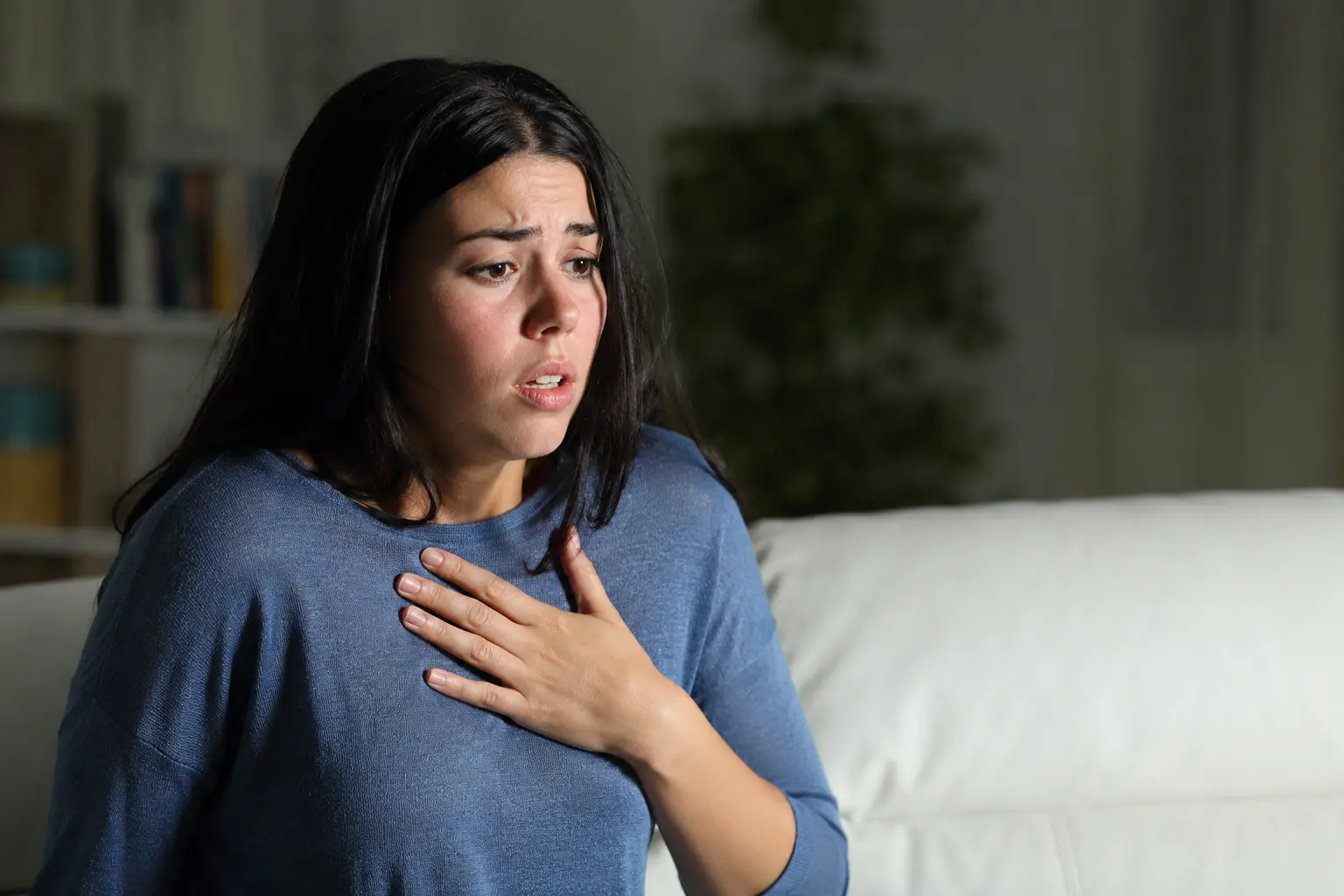 A woman experiencing a panic attack sits on a living room sofa with her hand on her chest, illustrating the symptoms of panic disorder in a realistic home setting 