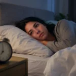 An anxious woman lying awake in bed at night, looking restless beside an alarm clock in a dimly lit bedroom 